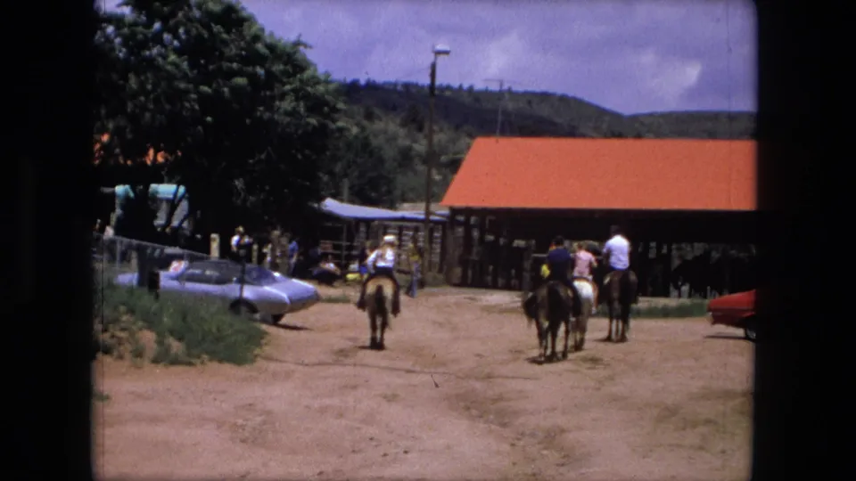 Home-movie frame from Fort Wayne in 1976 showing horseback riders in a sunny ranch yard with vintage cars nearby.