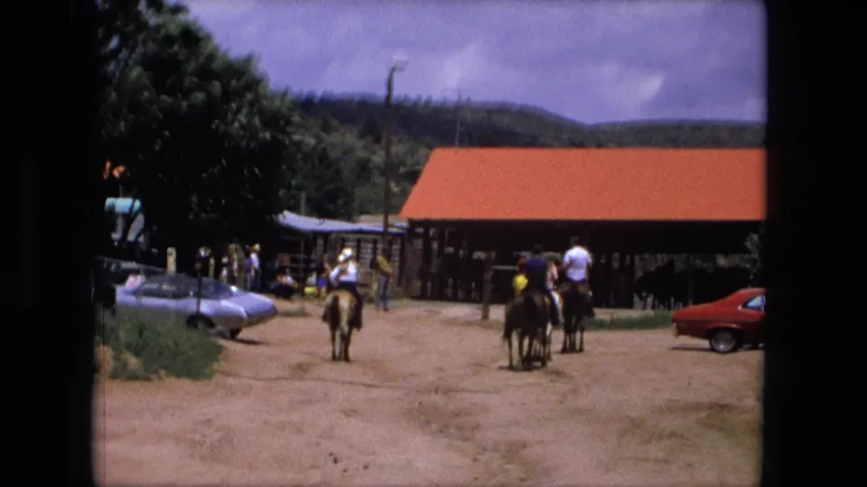 1976 Fort Wayne ranch footage frame with several riders on horseback crossing a dusty yard under a bright sky.