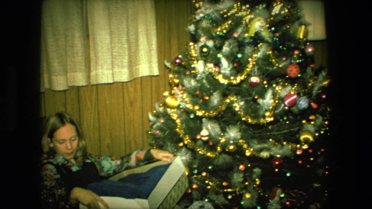 Woman opening a gift box beside a decorated Christmas tree in a wood-paneled room.