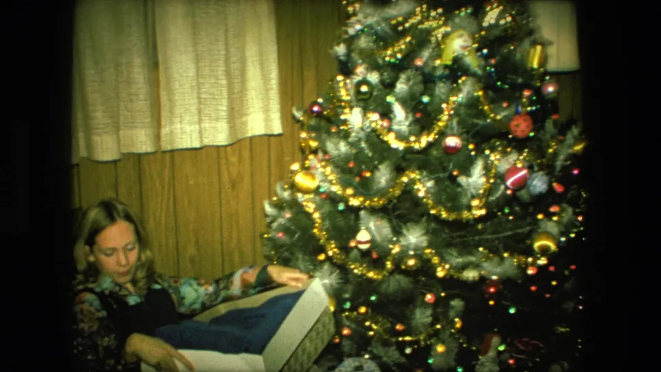 In 1976 Fort Wayne home-movie footage, a woman sits near a lit Christmas tree and looks into a gift box.