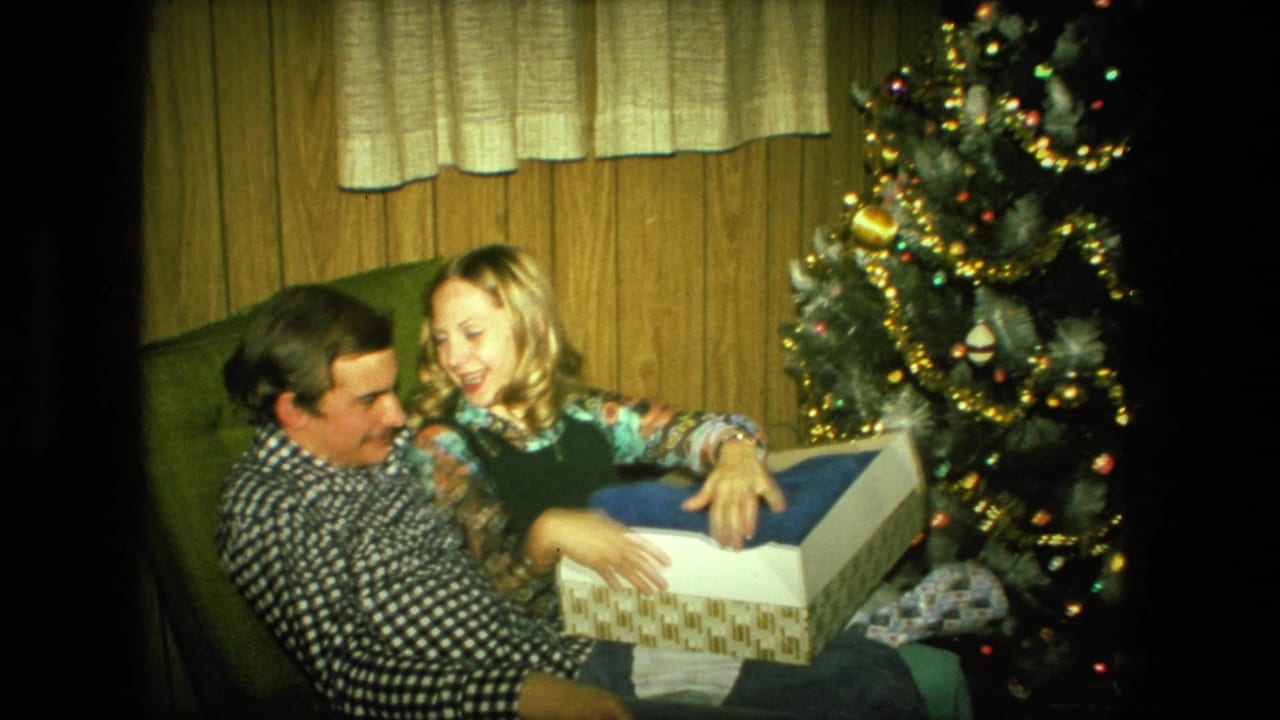 A man and woman sit closely on a green armchair in a Fort Wayne interior as she holds an opened Christmas gift box.