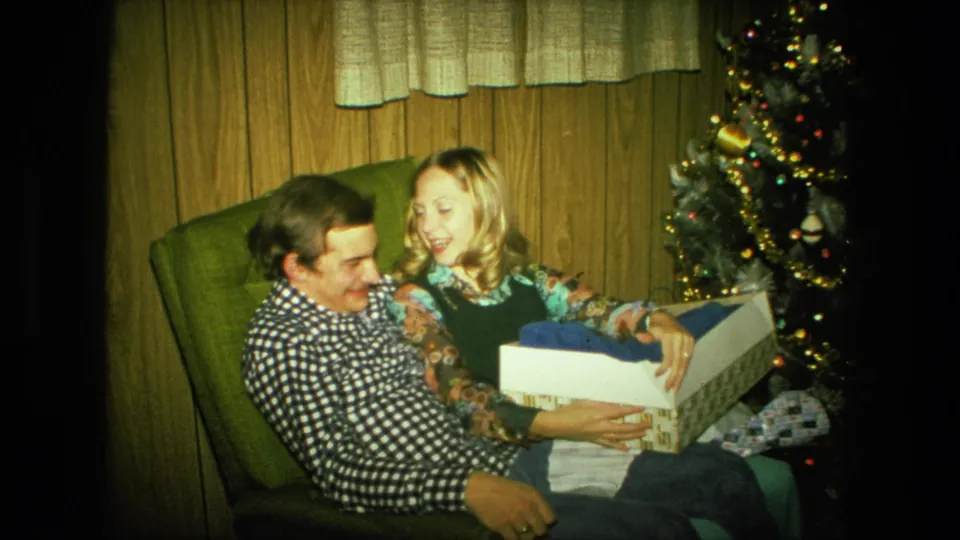 The couple sits in a green chair beside the lit tree, sharing a close Christmas gift-opening moment in 1976 home footage.