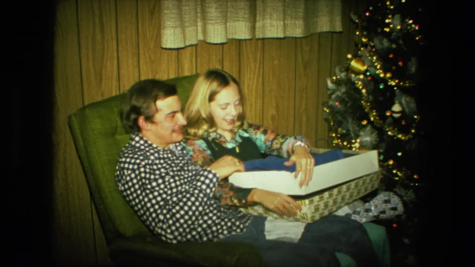 A smiling man and woman sit together by the Christmas tree, holding an open box in a wood-paneled Fort Wayne room.