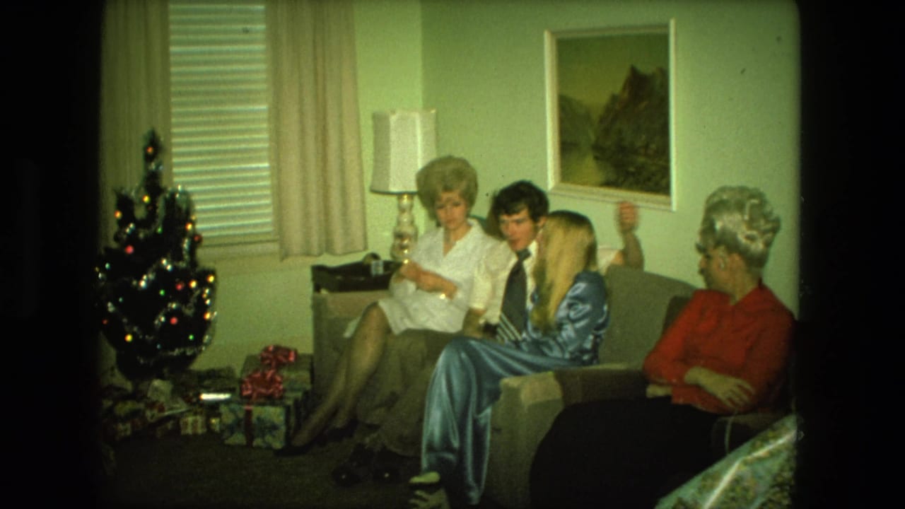 1976 Fort Wayne home-movie frame of four adults gathered on a living-room couch beside a small Christmas tree and presents.