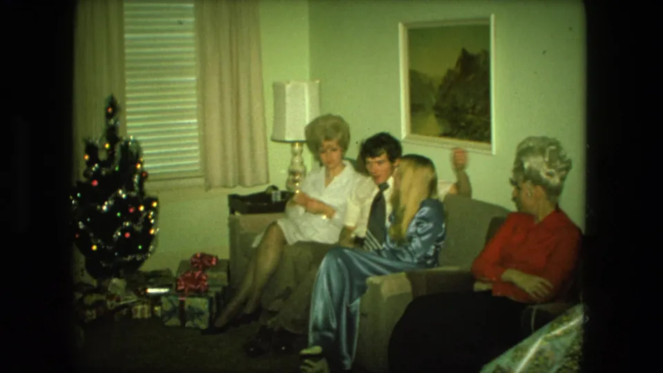 1976 Fort Wayne home-movie frame of four adults gathered on a living-room couch beside a small Christmas tree and presents.