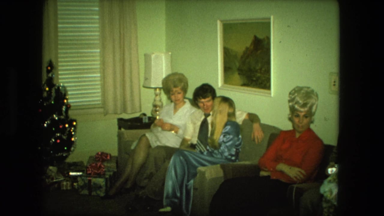 1976 Fort Wayne living-room frame with adults on a couch, a woman in red looking toward the camera, and Christmas decor at left.