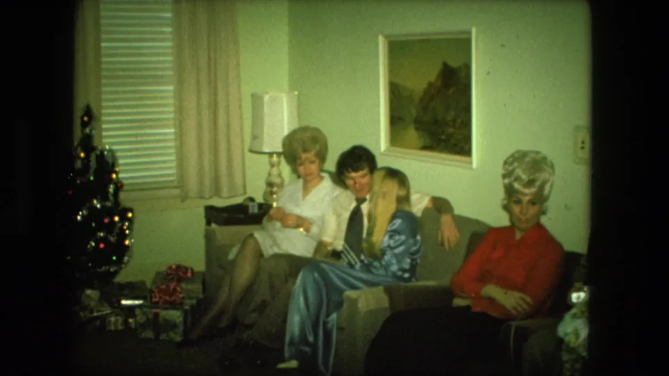 1976 Fort Wayne living-room frame with adults on a couch, a woman in red looking toward the camera, and Christmas decor at left.