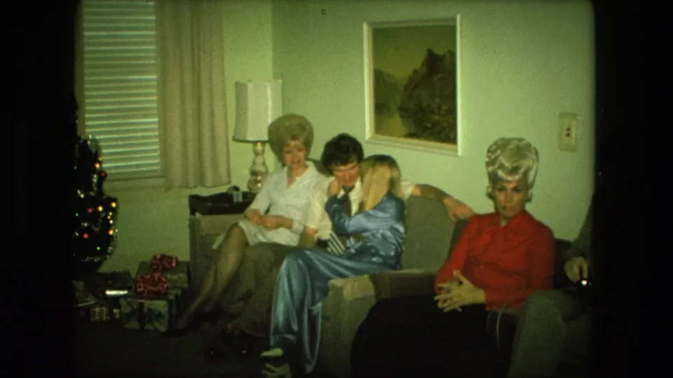 1976 Fort Wayne home-movie frame showing adults seated closely on a couch in a Christmas-decorated private living room.