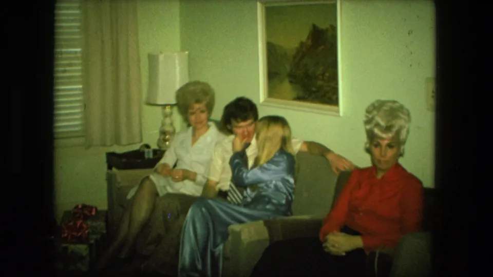 1976 Fort Wayne frame of seated adults in a holiday living room, with a man and woman leaning close near Christmas gifts and decor.