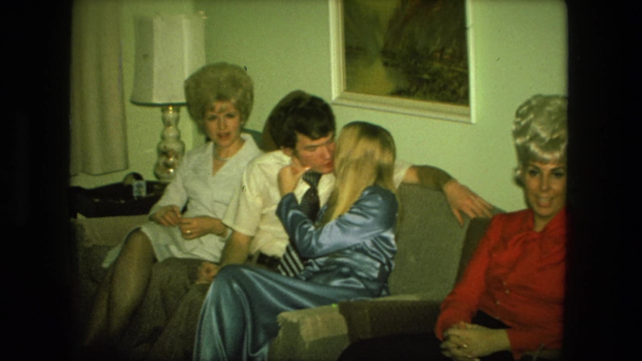 Adults sit on a living room couch as a couple kisses beside another woman.