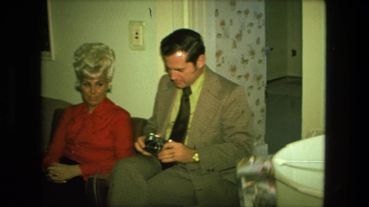 A woman in a red blouse sits next to a man in a patterned suit holding a camera in a private Fort Wayne living room.