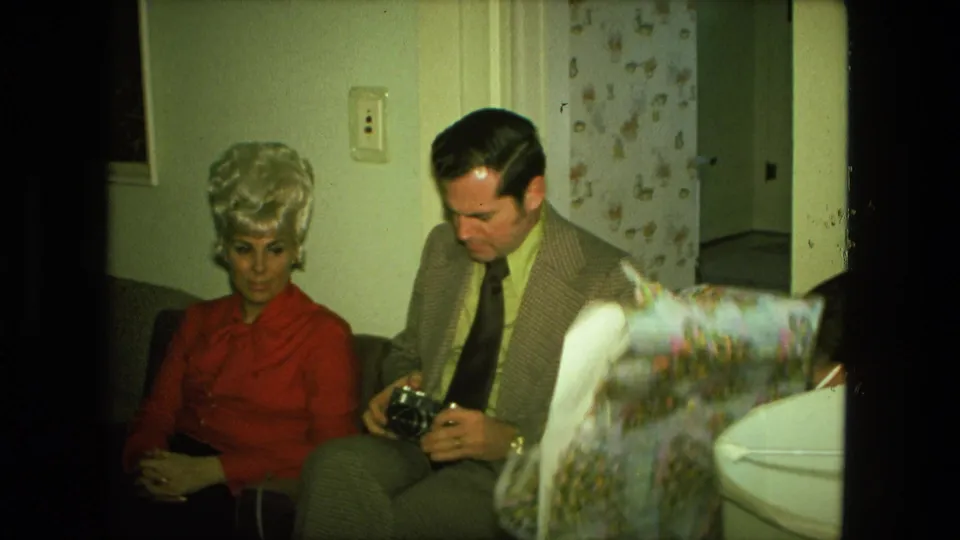 In the 1976 home-movie scene, a man studies a camera beside a seated woman as patterned holiday paper enters the frame.