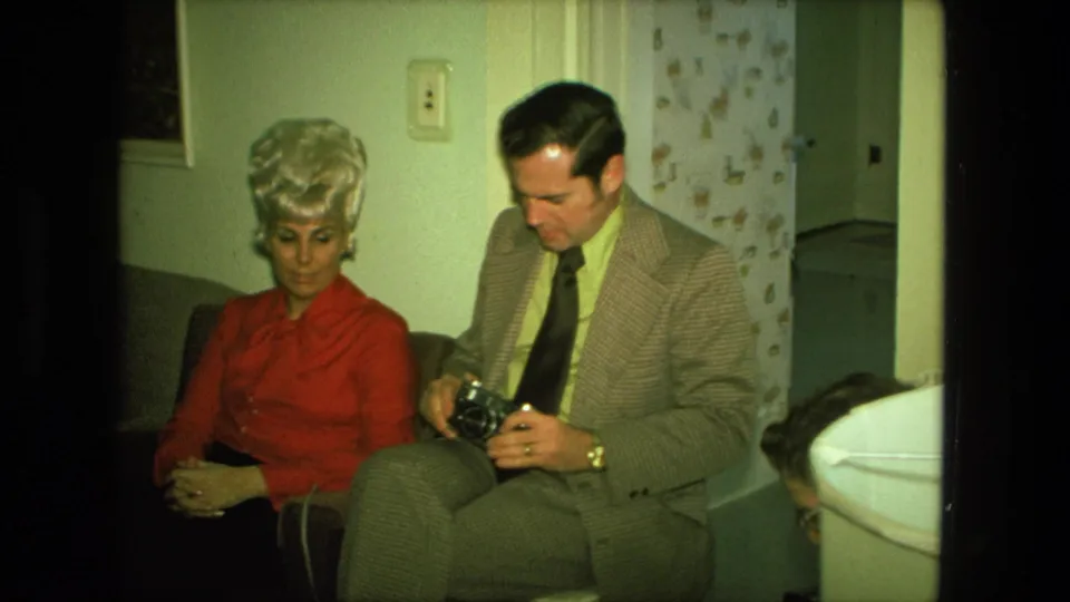 A man in a patterned suit looks down at a camera next to a woman in a red blouse in a 1970s private living room.