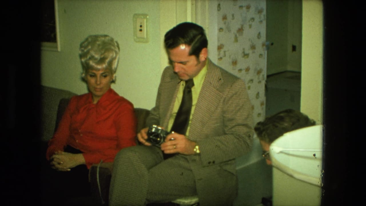 In a Fort Wayne 1976 home-movie frame, a blonde woman in red sits next to a man in a patterned suit examining a camera.