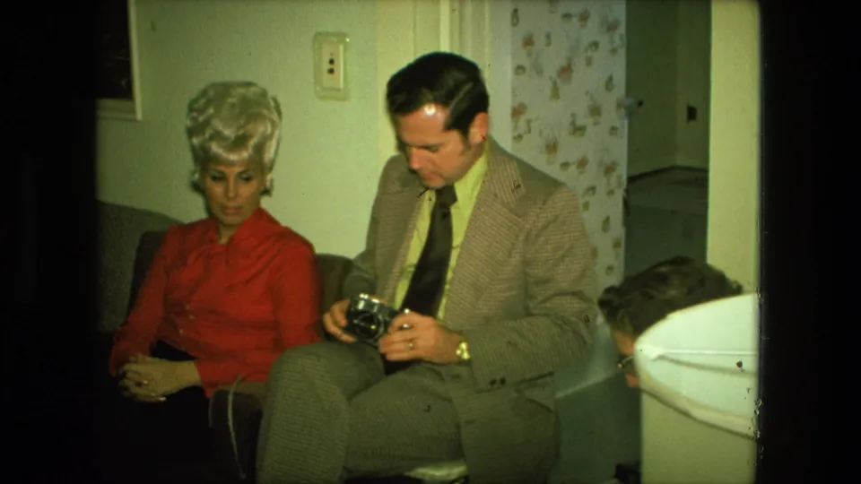 In a Fort Wayne 1976 home-movie frame, a blonde woman in red sits next to a man in a patterned suit examining a camera.