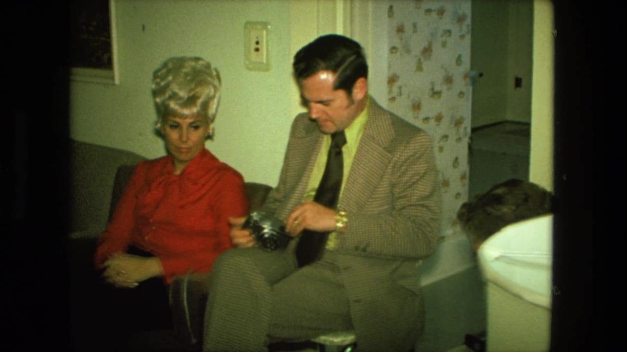 The man adjusts or inspects a camera while seated beside the blonde woman in a 1976 Fort Wayne private interior.