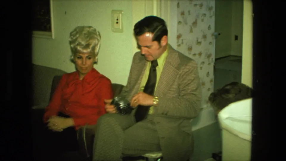 The man adjusts or inspects a camera while seated beside the blonde woman in a 1976 Fort Wayne private interior.