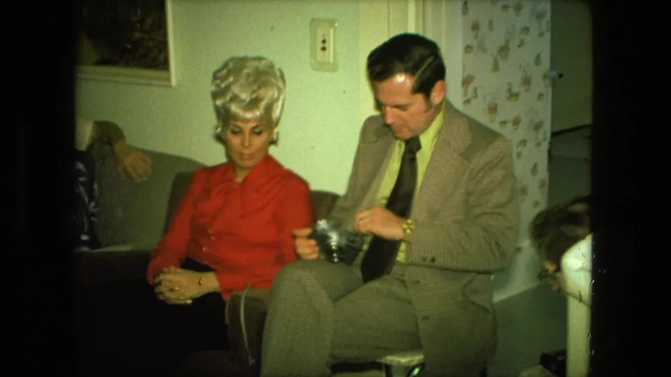 A 1970s home-movie frame shows the seated pair indoors, with the man handling a camera and the woman looking downward.