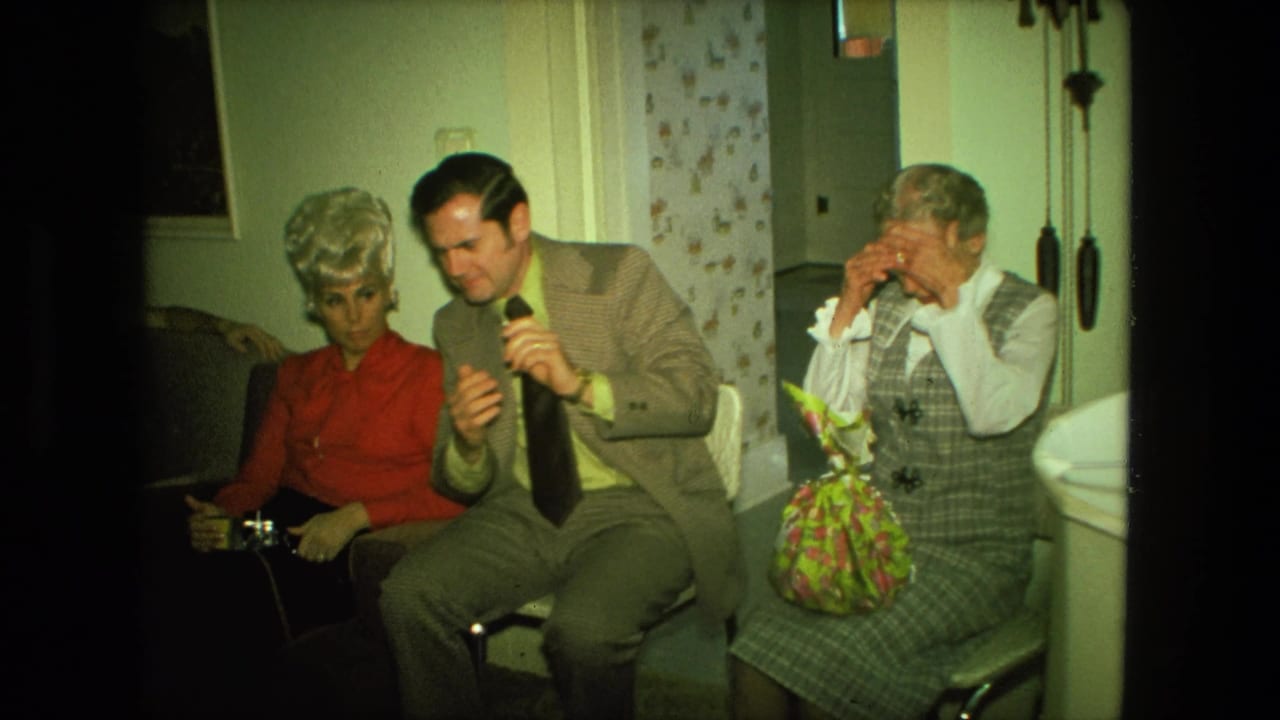 1976 Fort Wayne home-movie frame showing adults seated in a living room, with a suited man beside a woman in red and an older woman nearby.