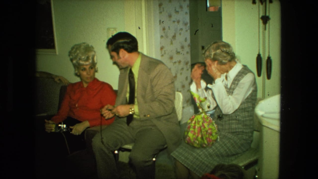 1976 Fort Wayne living-room scene with a suited man speaking to a woman in red as an older woman sits beside a floral gift bag.