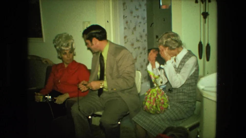 1976 Fort Wayne living-room scene with a suited man speaking to a woman in red as an older woman sits beside a floral gift bag.