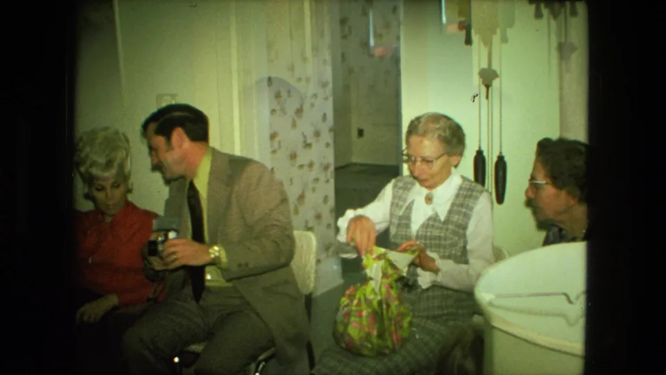 1976 Fort Wayne home-movie frame showing an older woman opening a floral gift bag while adults sit together in a private room.