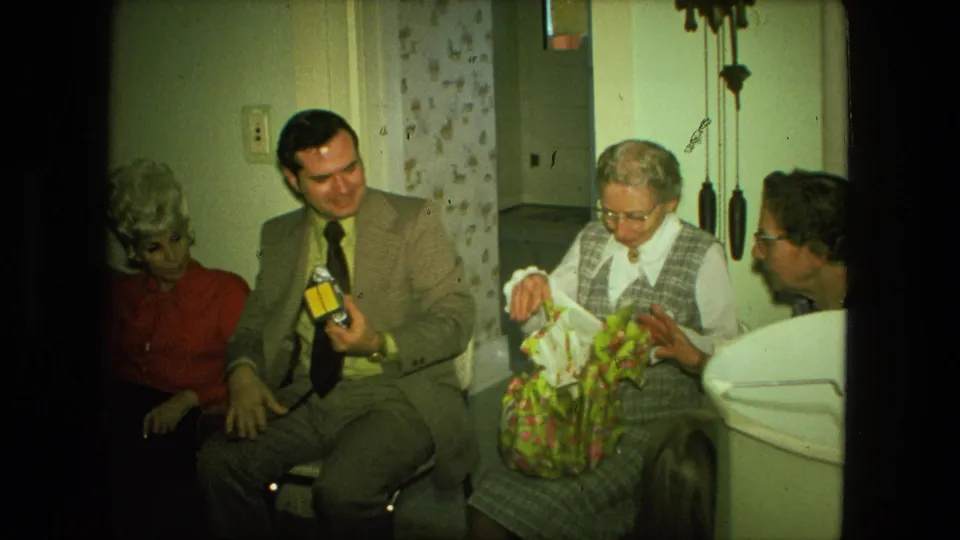 1976 Fort Wayne family gathering frame with an older woman opening a floral gift bag as seated adults watch in a living-room interior.