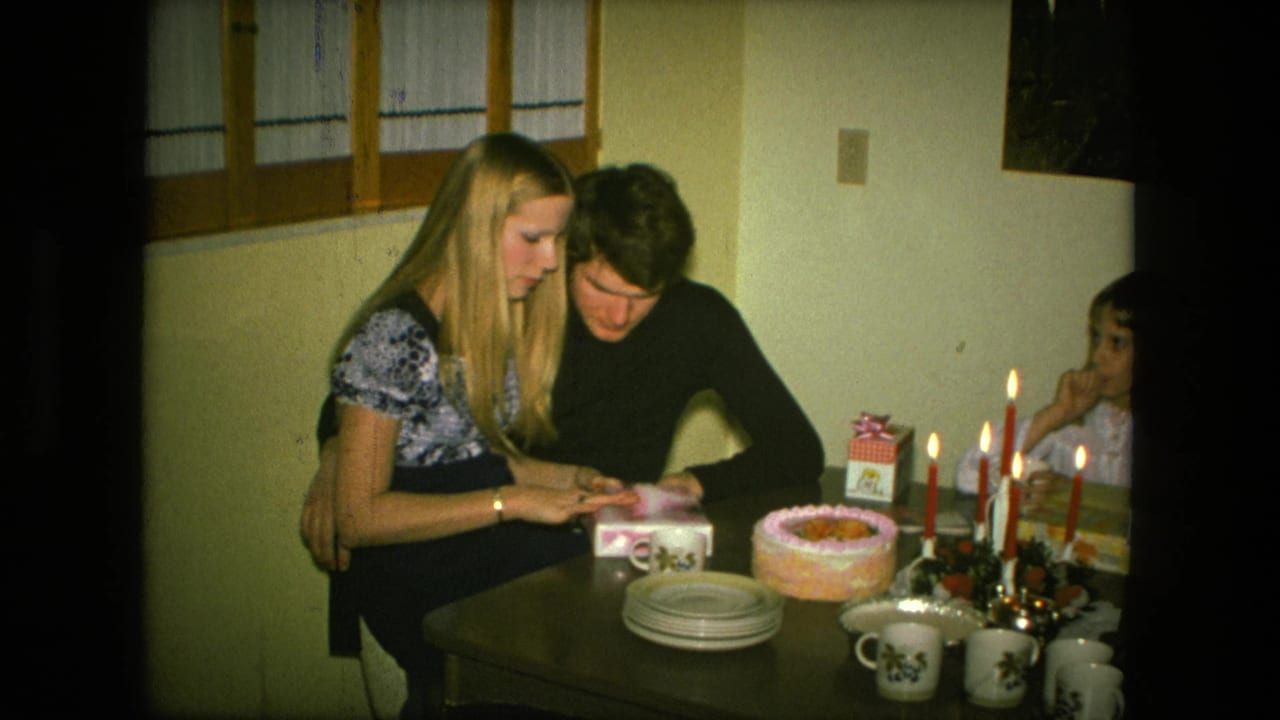 In Fort Wayne, Indiana in 1976, a young couple sits close at a birthday table as she handles a small gift beside cake and lit candles.