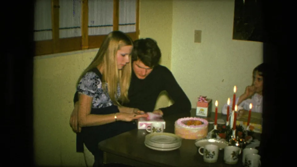 In Fort Wayne, Indiana in 1976, a young couple sits close at a birthday table as she handles a small gift beside cake and lit candles.