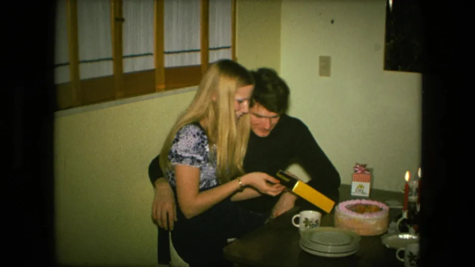 At the Fort Wayne birthday table, the couple leans together while opening a gift, with cake, plates, mugs, and candles in view.
