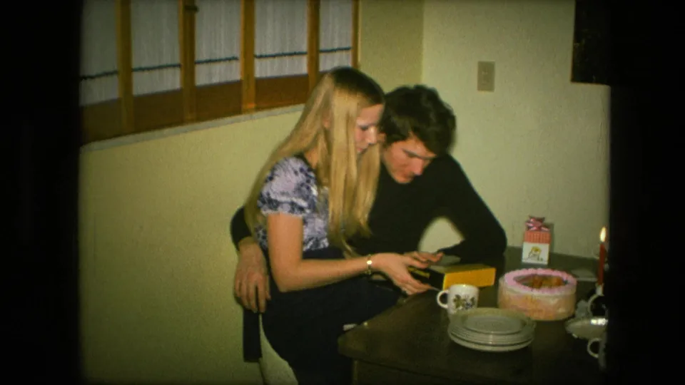 In the 1976 home-movie frame, the couple sits close and examines a small gift item beside a cake and candle arrangement.