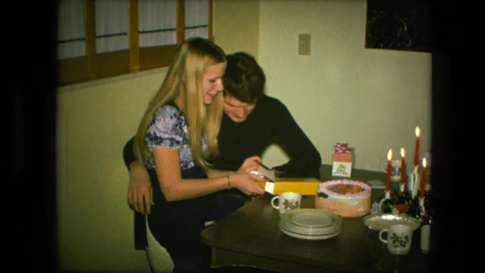 A blonde woman and young man focus on the gift box during a Fort Wayne birthday gathering filmed on 8mm in 1976.