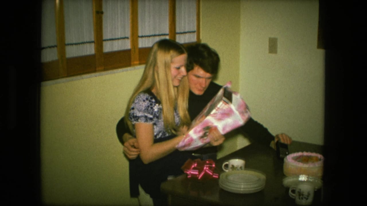 Young woman seated on a man's lap as they open pink wrapping paper at a dining table.