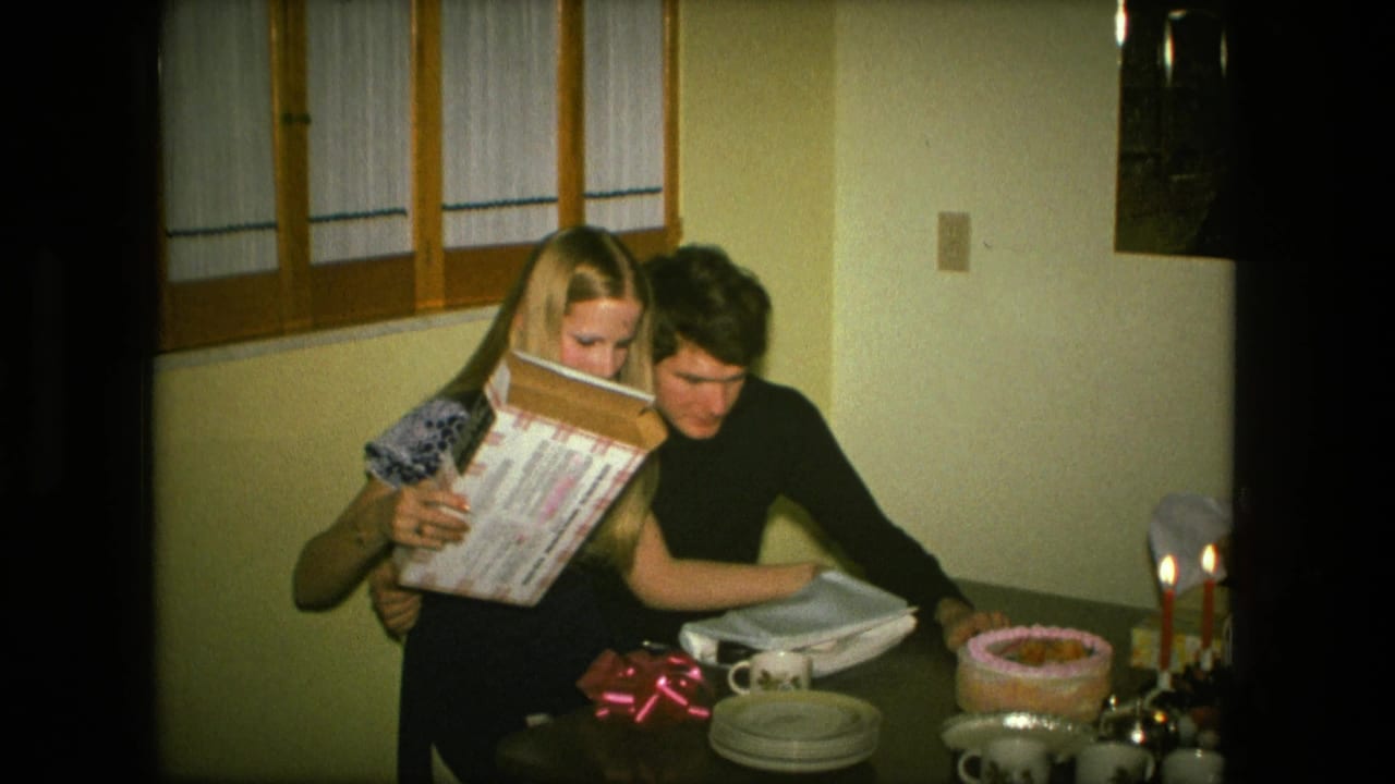 The couple looks over the opened gift together in a dim 1976 Fort Wayne dining room with cake and candles nearby.