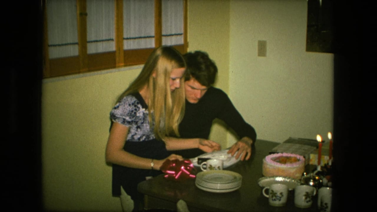 Young couple seated at a table opening a gift beside a birthday cake and lit candles.