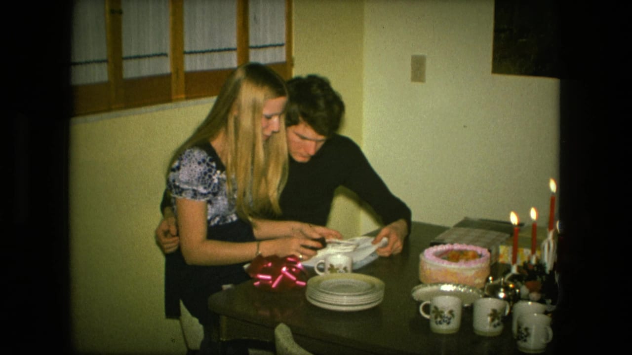 The couple remains seated closely at the Fort Wayne birthday table, handling wrapping paper as candles burn beside the cake.