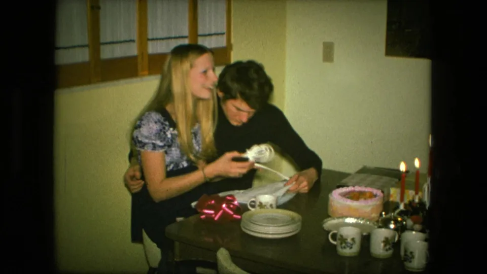 During the 1976 birthday scene, the woman lifts part of the opened present or wrapping while the man looks down at the table.