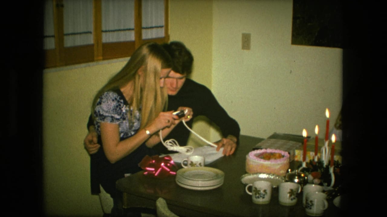 The woman examines a small item while the man leans beside her at a 1976 Fort Wayne birthday table with cake, candles, mugs, and plates.
