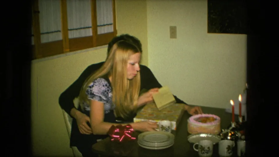 In the Fort Wayne home interior, the woman opens a wrapped present while the man holds her close beside a cake, candles, mugs, and plates.