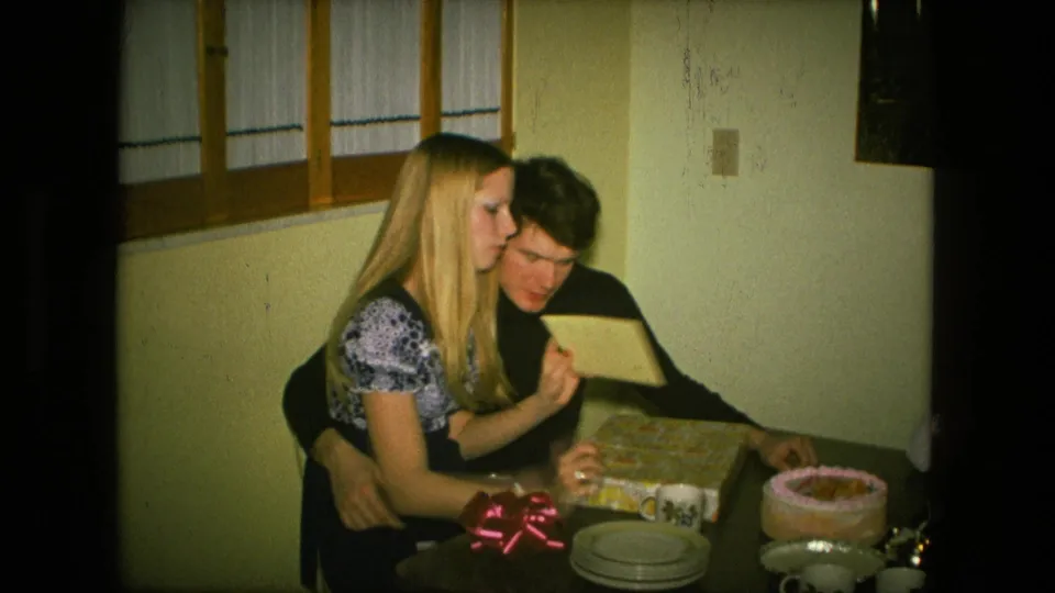 The couple reviews an opened card or paper at the dining table during the 1976 Fort Wayne birthday gathering, with cake and candles nearby.