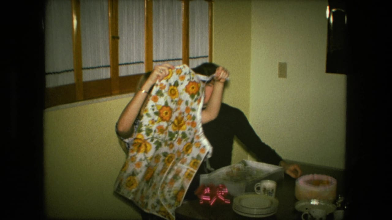 In a 1976 Fort Wayne home interior, a young woman raises a sunflower-patterned apron while a man sits beside an open gift box.
