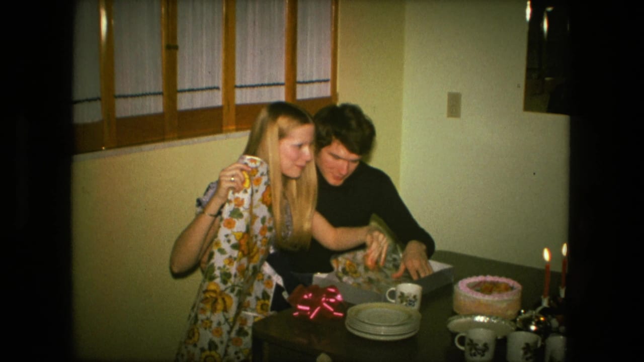 The couple leans together over an open gift box in a warm 1976 Fort Wayne dining room with cake, plates, cups, and candles nearby.