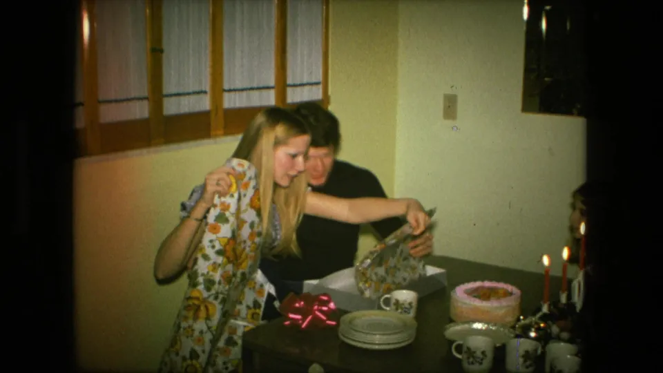 A young woman in a Fort Wayne home celebration holds a floral apron and reaches toward wrapping paper as her companion watches from behind.