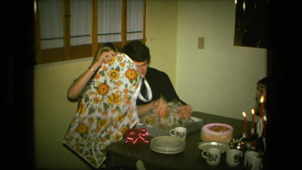 The apron is raised again as the man looks down at wrapping paper beside a party table with cake, cups, plates, candles, and an open box.