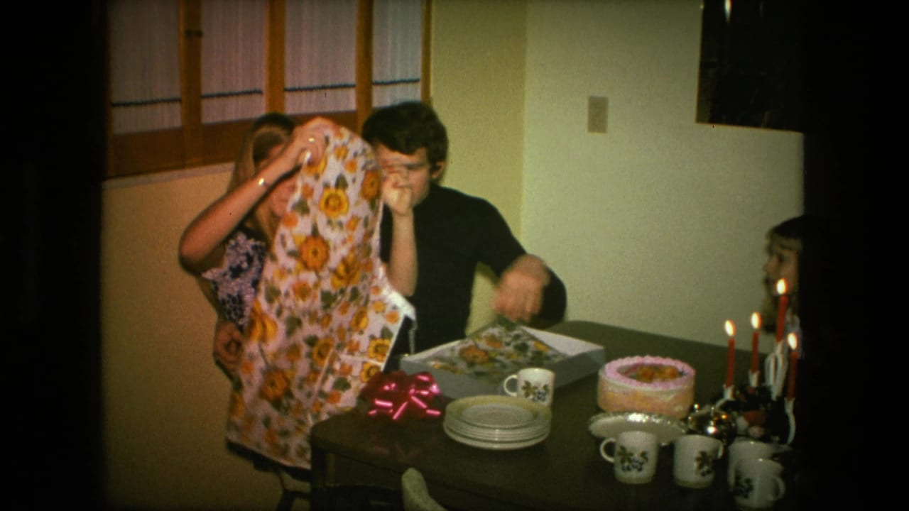 Woman lifts a sunflower apron while a man sits beside her at a candlelit birthday table.