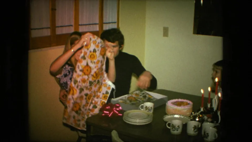 In a 1976 Fort Wayne home interior, a woman holds up a sunflower apron gift beside a man at a table with cake, candles, plates, and mugs.