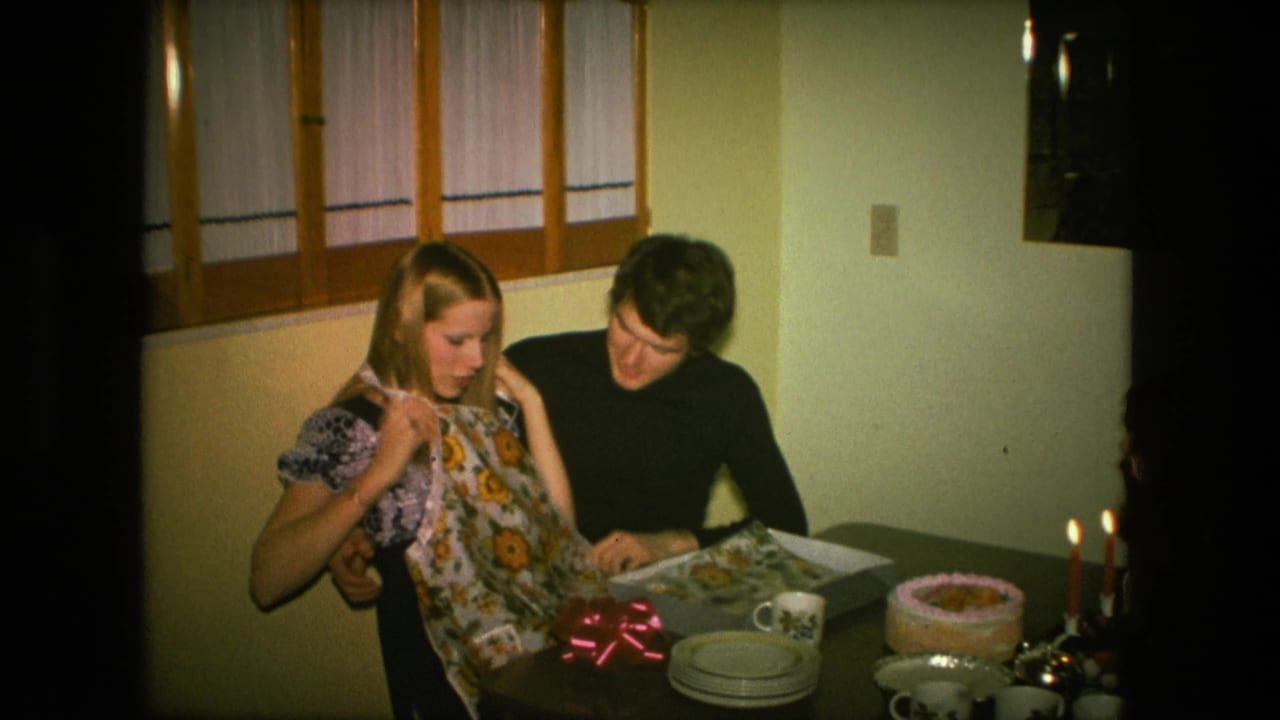 The couple looks down as the woman holds the sunflower apron in a Fort Wayne dining room, with a cake and lit candles on the table.