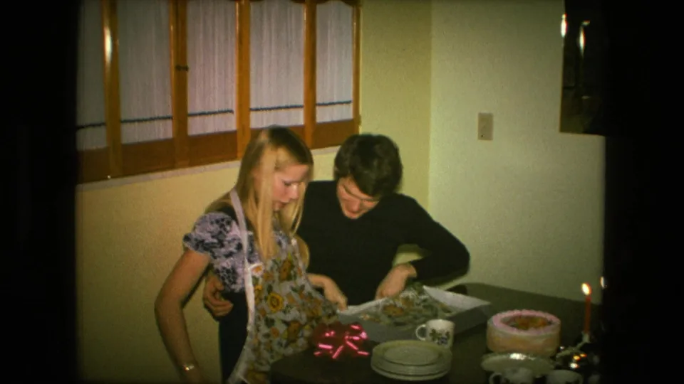 In the 1976 home-movie frame, the woman tries on the sunflower apron as the man holds her from behind beside cake, candles, plates, and mugs.