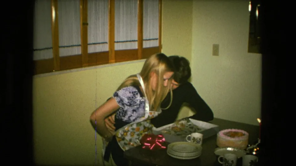 The woman and man lean together over the apron in a Fort Wayne dining room, with the cake, candles, stacked plates, and mugs still visible.