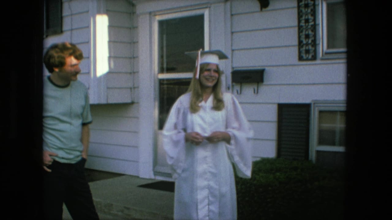 Graduate in white cap and gown stands outside a house with a young man beside her.